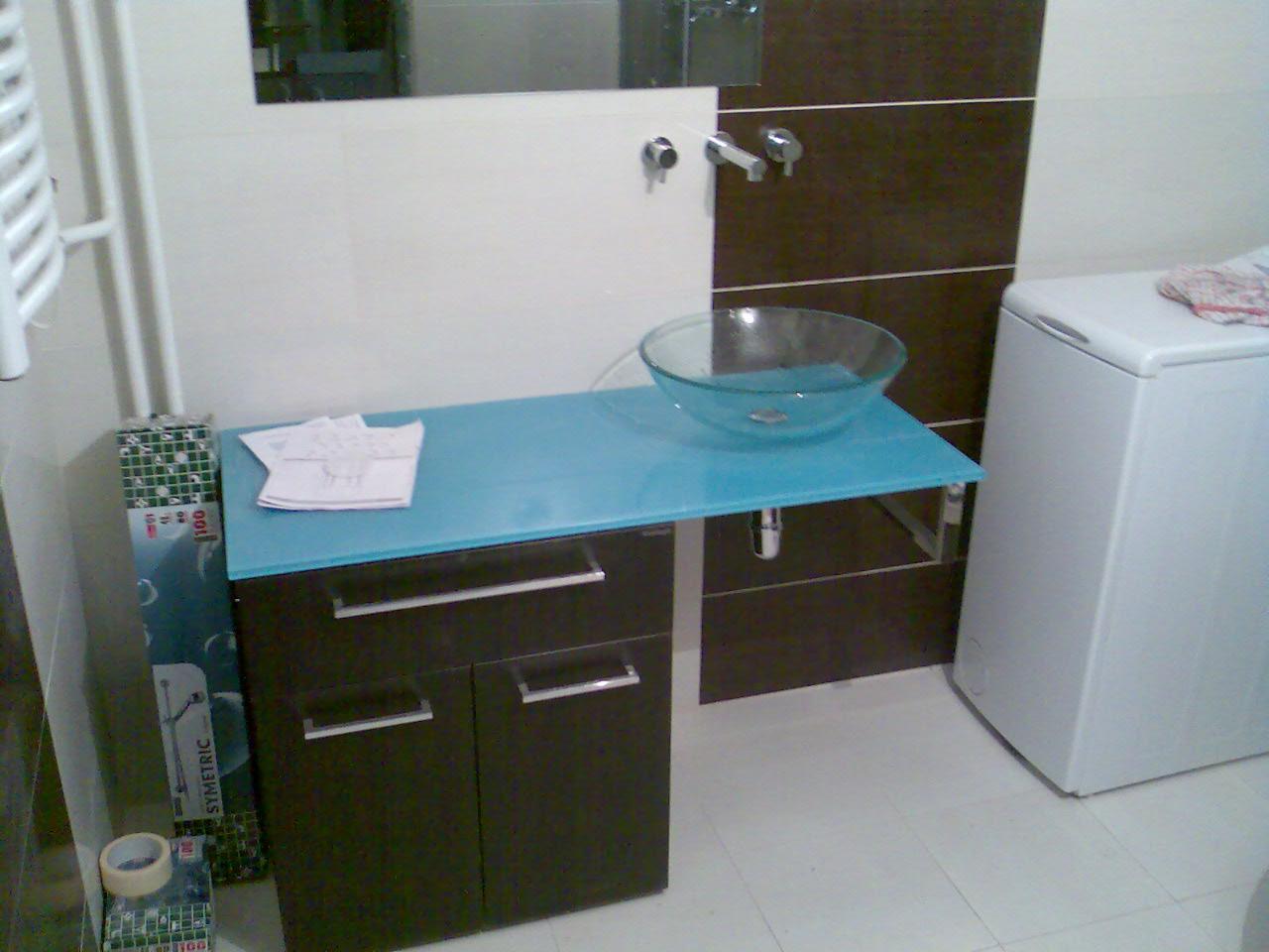 Łazienka z umywalką szklaną na niebieskim blacie, ciemna szafka pod umywalką, pralka z boku, płytki na ścianie w dwóch kolorach, widok na baterię i lustro.