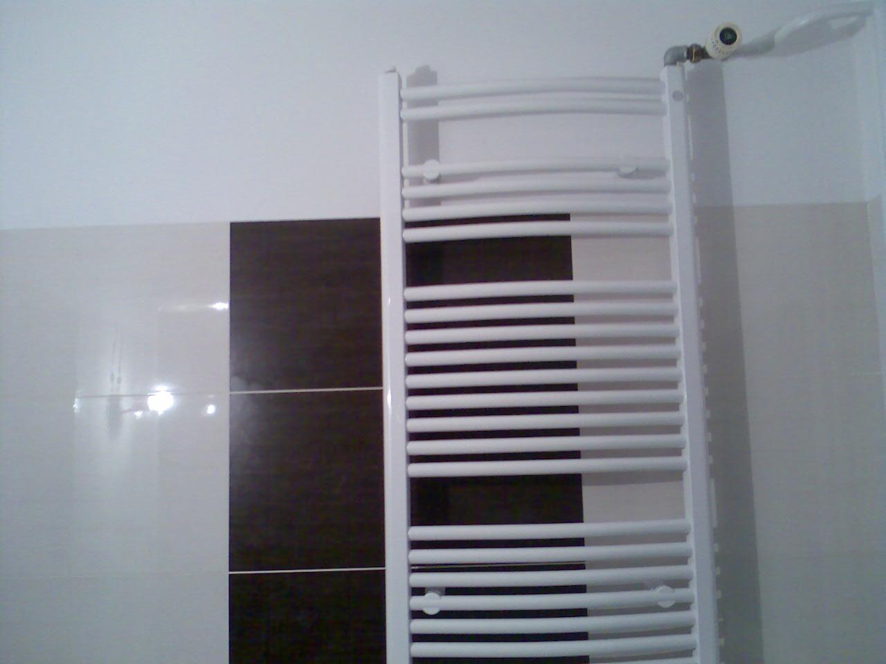 Biały grzejnik drabinkowy zamontowany na ścianie z białymi i ciemnobrązowymi płytkami, widoczny zawór termostatyczny.