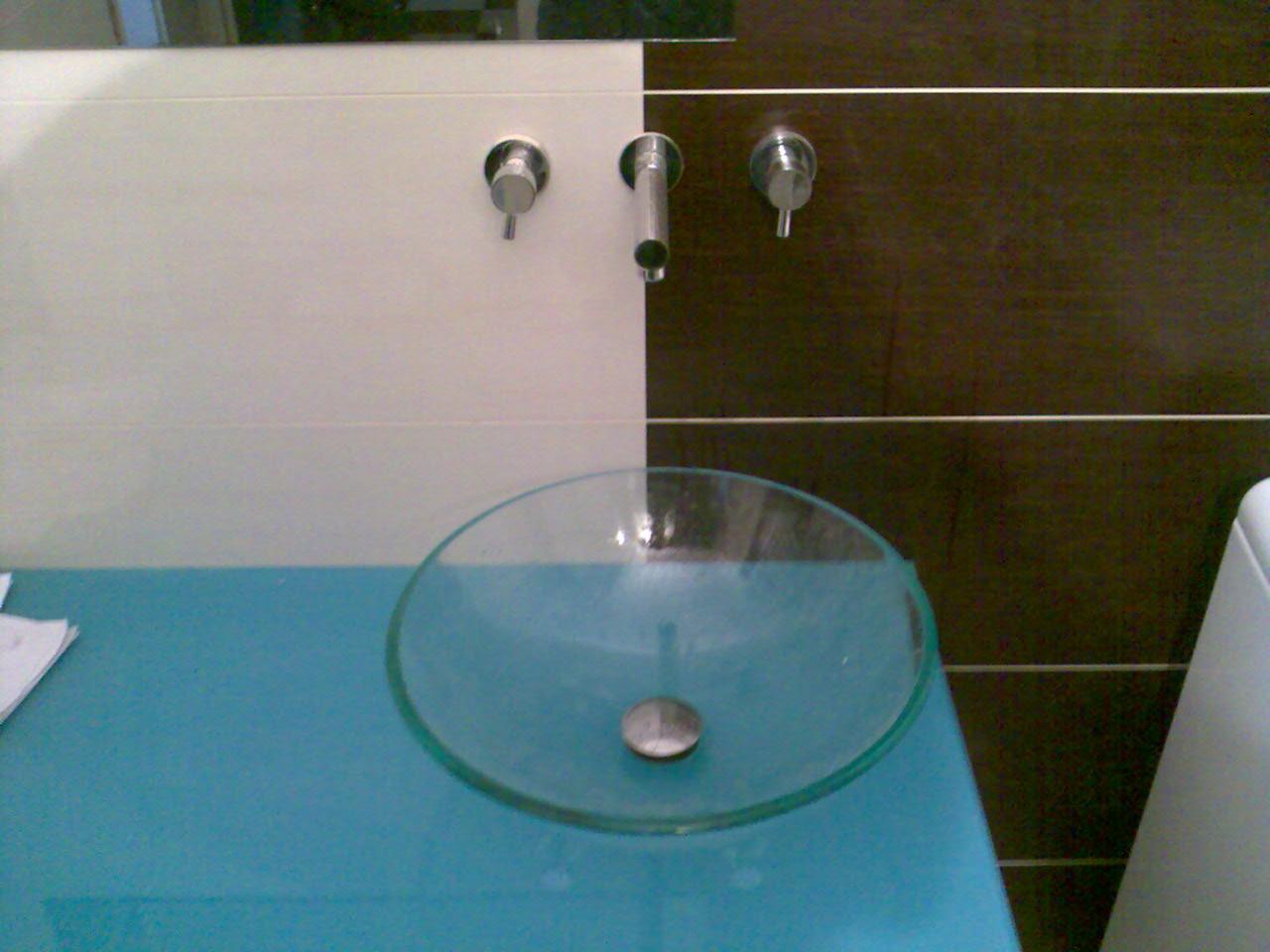 Szklana umywalka na turkusowym blacie w nowoczesnej łazience z białymi i brązowymi płytkami, trzy chromowane krany na ścianie.