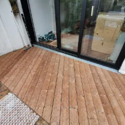 Drewniany taras z desek, częściowo ułożony, z widoczną konstrukcją pod spodem, narzędzia akumulatorowe Makita widoczne przez przesuwne drzwi tarasowe.