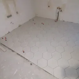 Układanie jasnoszarych, heksagonalnych płytek podłogowych w łazience, z użyciem czerwonych klinów poziomujących, widoczne surowe ściany i wystające rury instalacji wodnej.