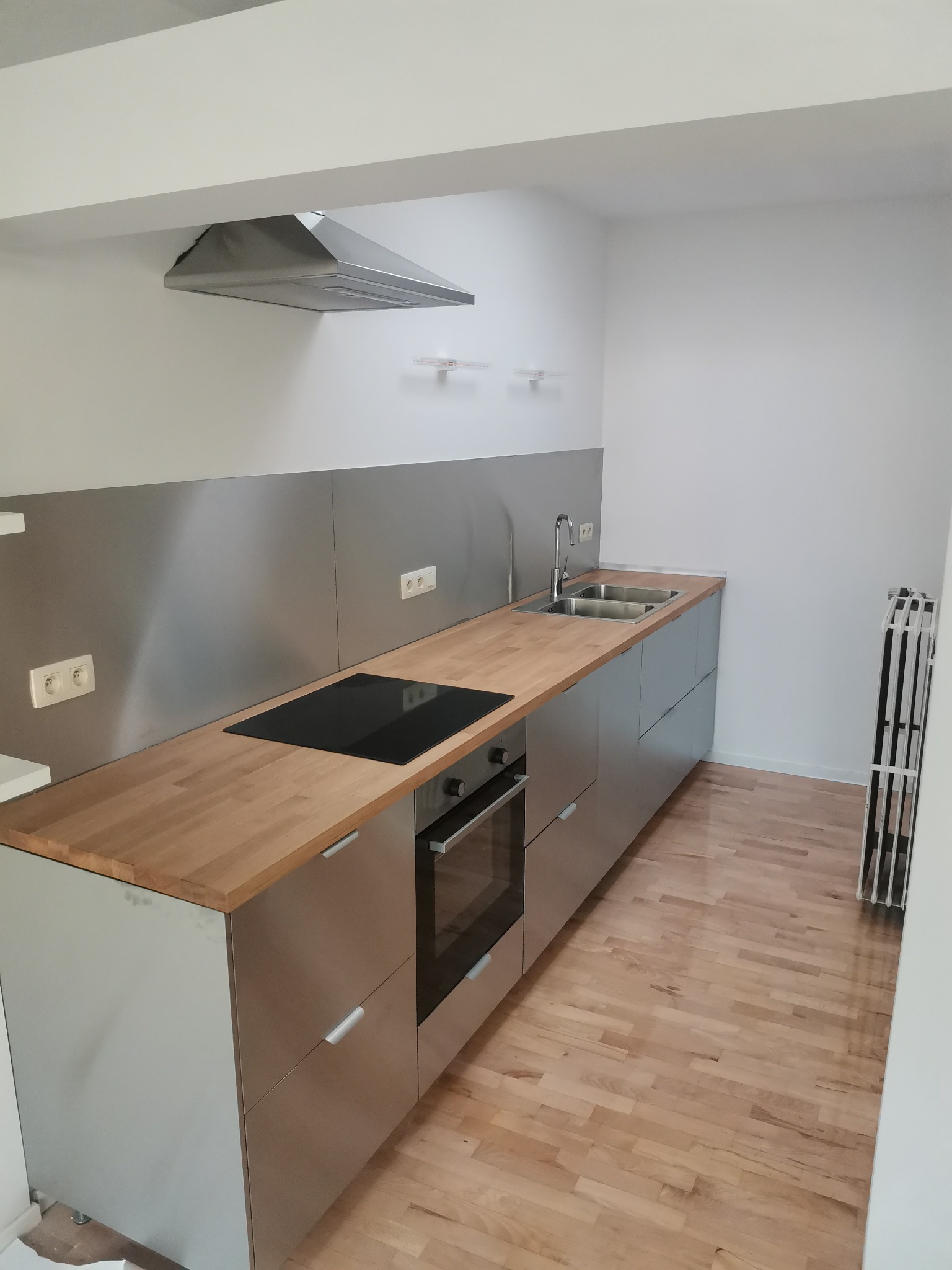 Nowoczesna kuchnia z szarymi szafkami, drewnianym blatem, płytą indukcyjną i stalowym panelem nad blatem. Na podłodze parkiet. Widoczny również stalowy okap i suszarka na pranie.