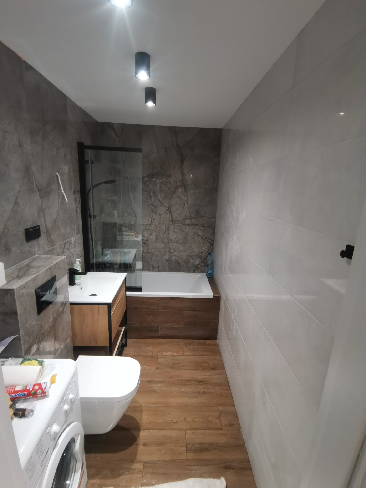 Nowoczesna łazienka po remoncie:  płytki imitujące marmur, drewnopodobna podłoga,  biała armatura, kabina prysznicowa z czarnymi ramami, pralka z widocznymi materiałami budowlanymi na wierzchu.