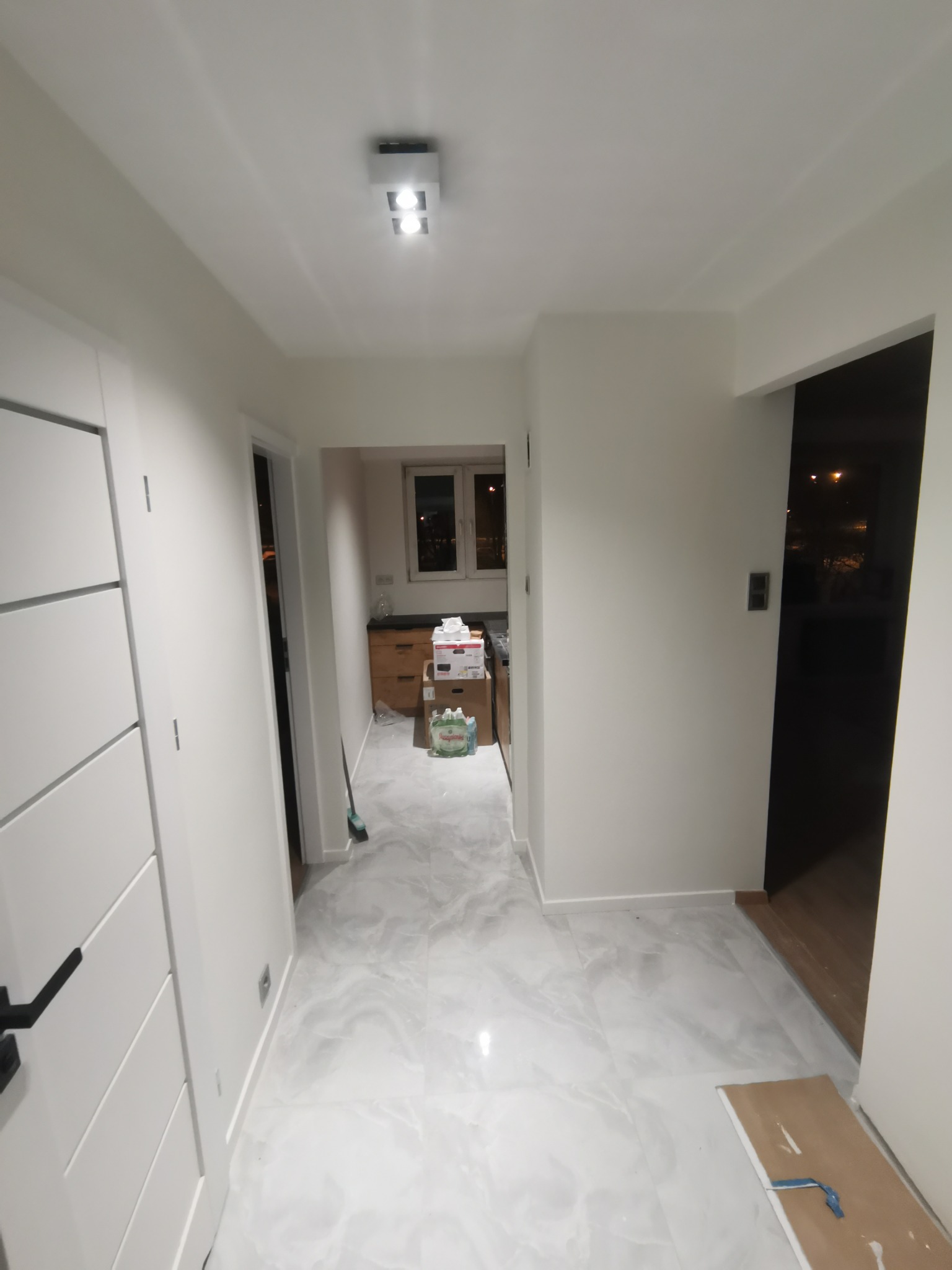 Korytarz po odświeżeniu z białymi ścianami, marmurową podłogą i białymi drzwiami, widoczne kartony w głębi pomieszczenia sugerujące trwające prace.