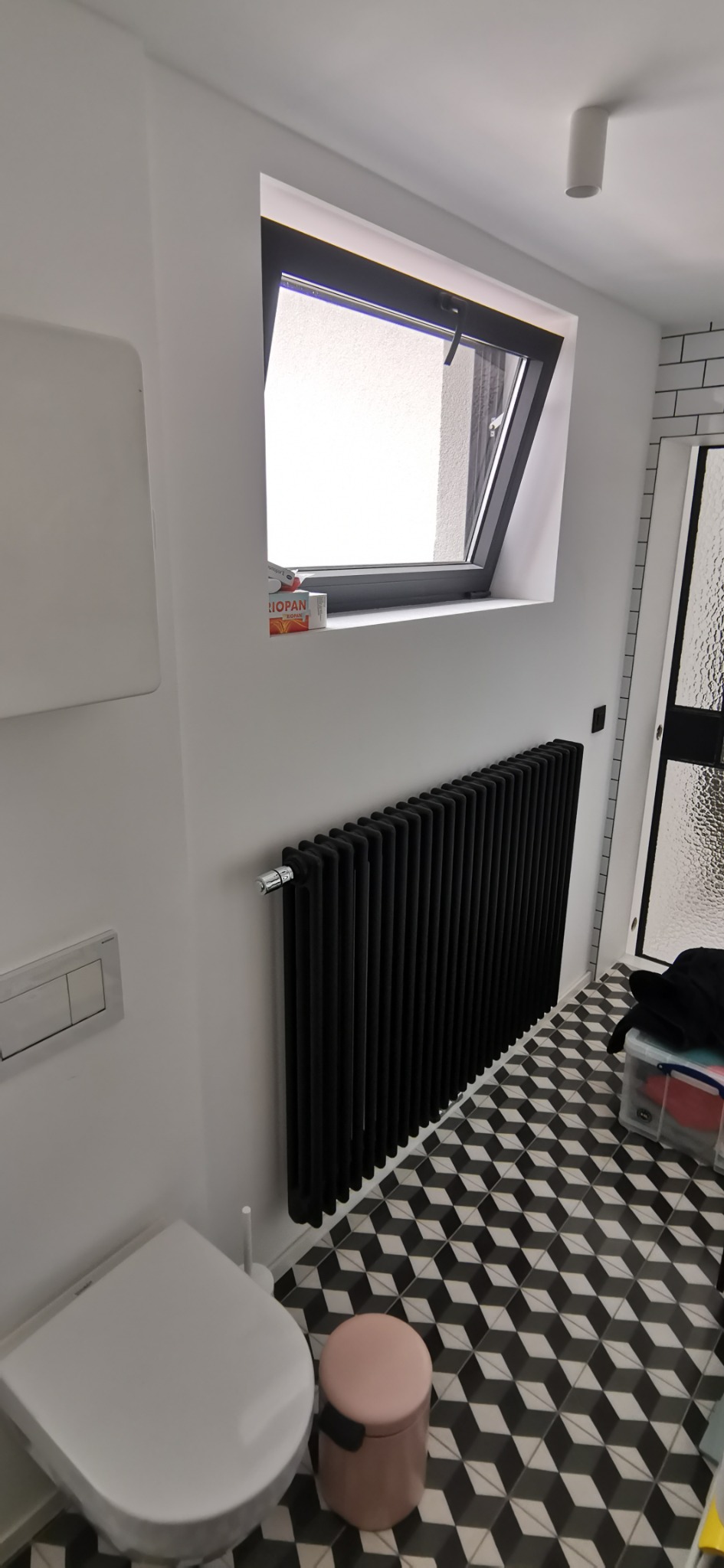 Biała łazienka z czarnym, żeliwnym grzejnikiem, wzorzystą podłogą i oknem dachowym, na parapecie pudełko leku.