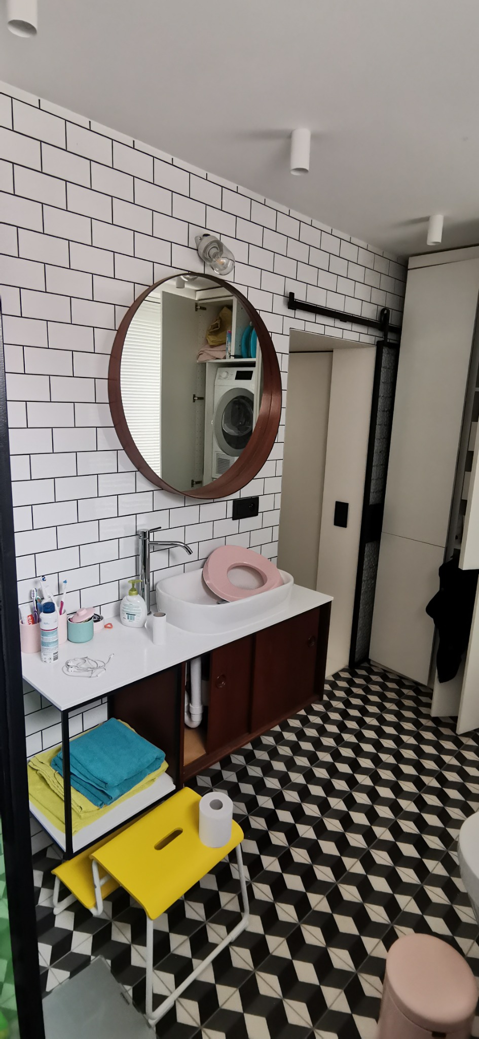 Umywalka nablatowa z różową deską sedesową w nowoczesnej łazience z białymi płytkami i czarno-białą podłogą w romby, widoczne żółte taborety i pralka w lustrze.