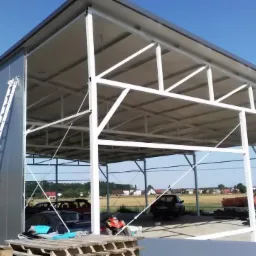 Gotowa hala stalowa z szarymi ścianami i białym szkieletem konstrukcyjnym, widoczna drabina oparta o ścianę, wewnątrz zaparkowane pojazdy i palety na podłodze, w tle krajobraz wiejski z polami...