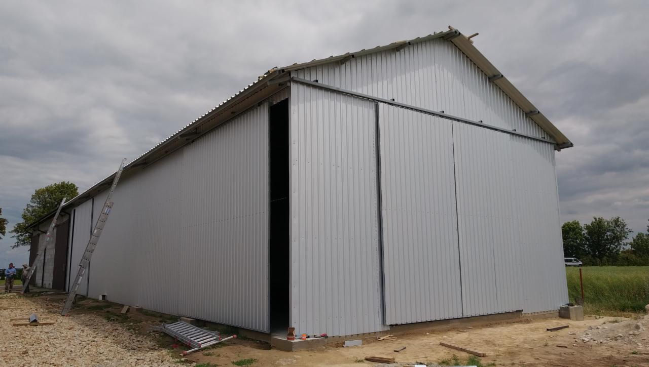 Budowa hali z płyt warstwowych w trakcie montażu, widoczne otwarte wrota przesuwne, drabina oparta o ścianę, teren budowy.