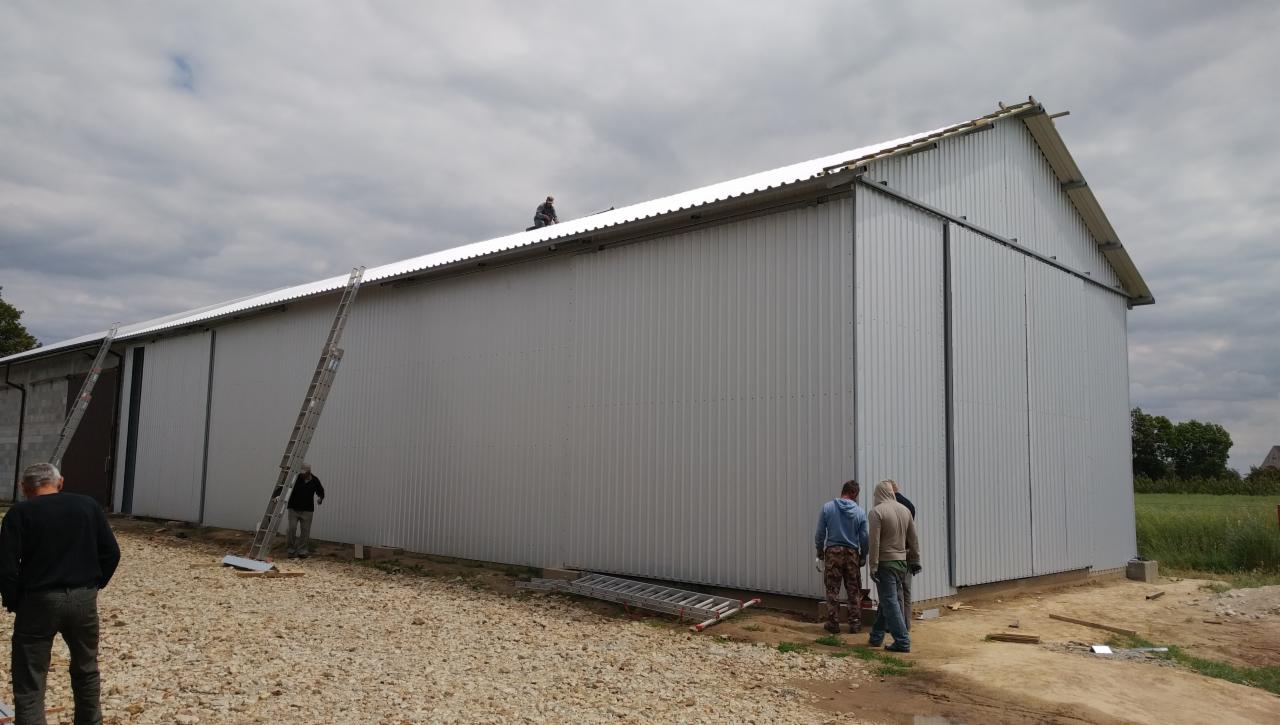 Montaż hali z płyt warstwowych w trakcie realizacji, widoczni pracownicy na dachu i przy ścianach, drabiny opierają się o budynek, teren wokół wysypany żwirem, pochmurne niebo.