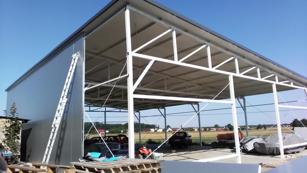 Gotowa hala stalowa z szarymi ścianami i białym szkieletem konstrukcyjnym, widoczna drabina oparta o ścianę, wewnątrz zaparkowane pojazdy i palety na podłodze, w tle krajobraz wiejski z polami...