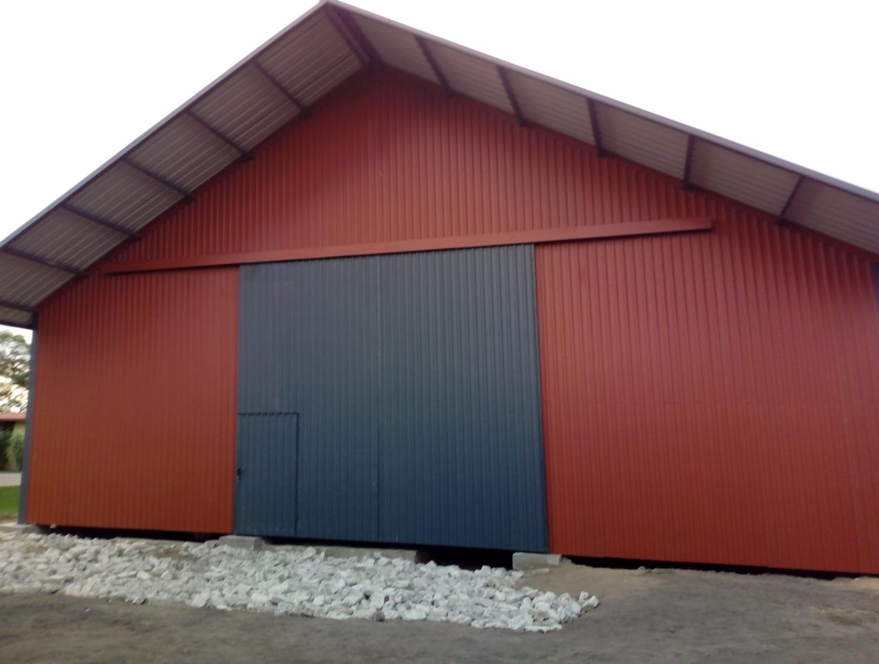 Czerwono-szara hala z przesuwanymi drzwiami i małym wejściem, wykończona kamieniami u podstawy, widok z zewnątrz.