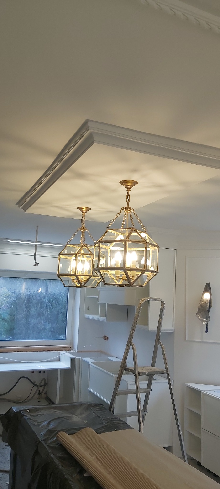 Dwa złote, geometryczne żyrandole zwisające z sufitu z ozdobną sztukaterią w remontowanej kuchni, widoczna drabina i zabezpieczone folią meble.