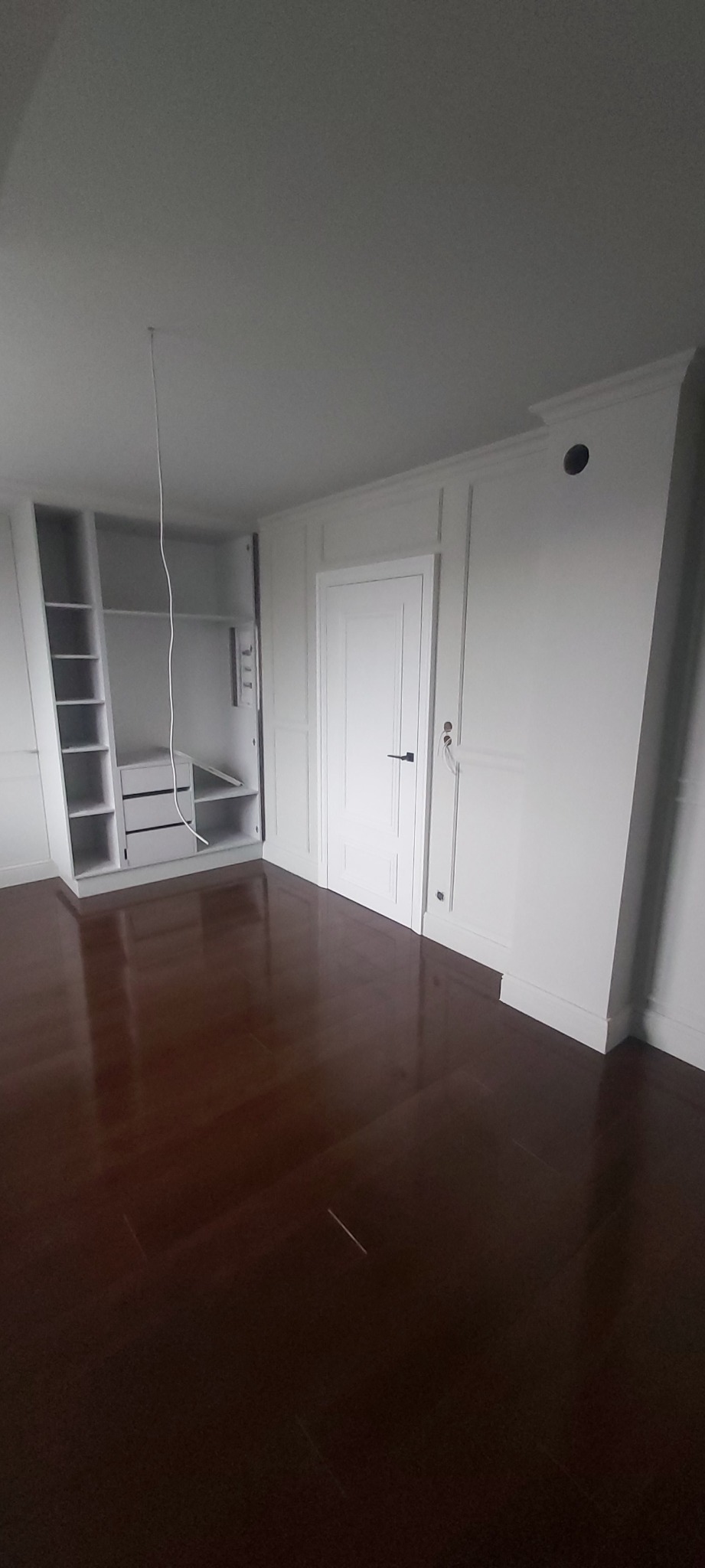 Białe wnętrze pokoju z otwartą szafą wnękową z półkami i szufladami, ciemna drewniana podłoga, białe ściany z dekoracyjnymi panelami.