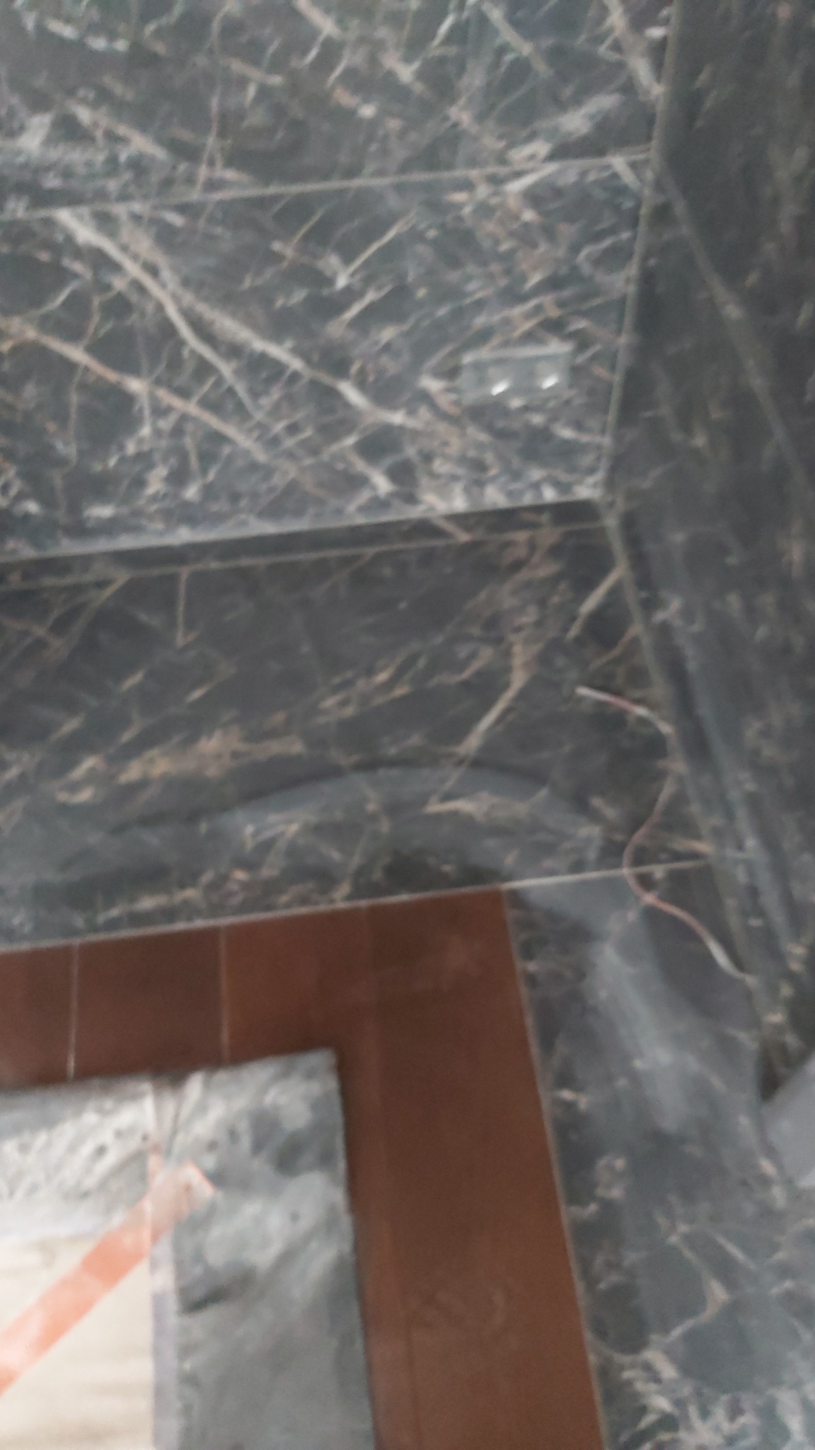 Fragment ściany i podłogi wyłożonej płytkami imitującymi marmur, z widocznymi brązowymi płytkami podłogowymi i wystającym kablem elektrycznym.