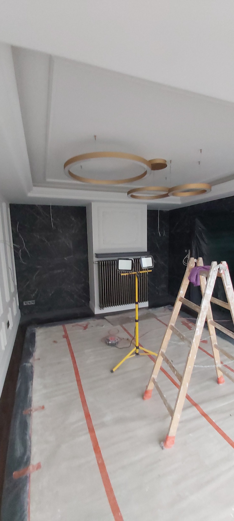 Wnętrze w trakcie prac wykończeniowych: zabezpieczona folią podłoga z taśmą, drabina, lampa budowlana, grzejnik pod dekoracyjną ramą i marmurowa ściana pod sufitem z okrągłymi lampami.