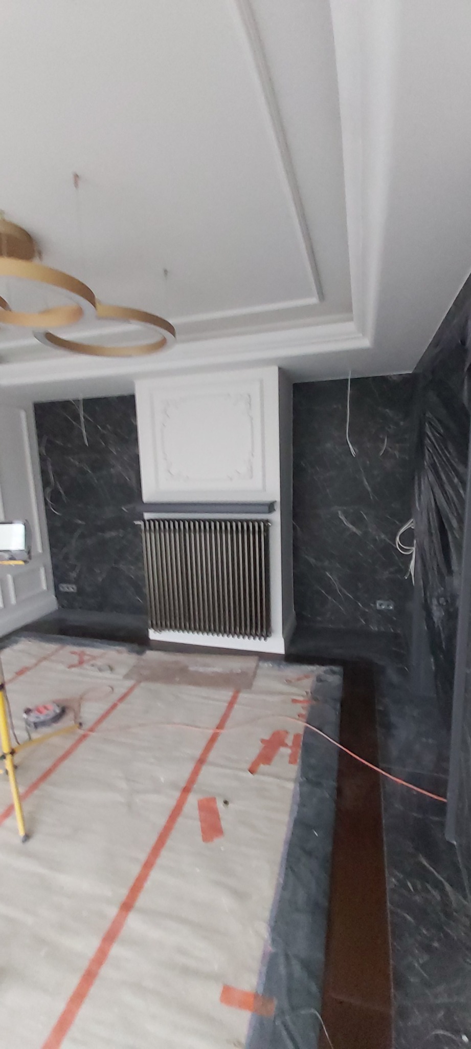 Wnętrze pokoju w trakcie prac wykończeniowych: marmurowe ściany, grzejnik w obudowie z dekoracyjną sztukaterią, zabezpieczona folią podłoga i wiszące kable elektryczne.