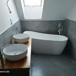 Nowoczesna łazienka na poddaszu z dwoma umywalkami nablatowymi na drewnianym blacie, wolnostojącą wanną i szarą glazurą, widoczne niedokończone podłączenie elektryczne na ścianie.