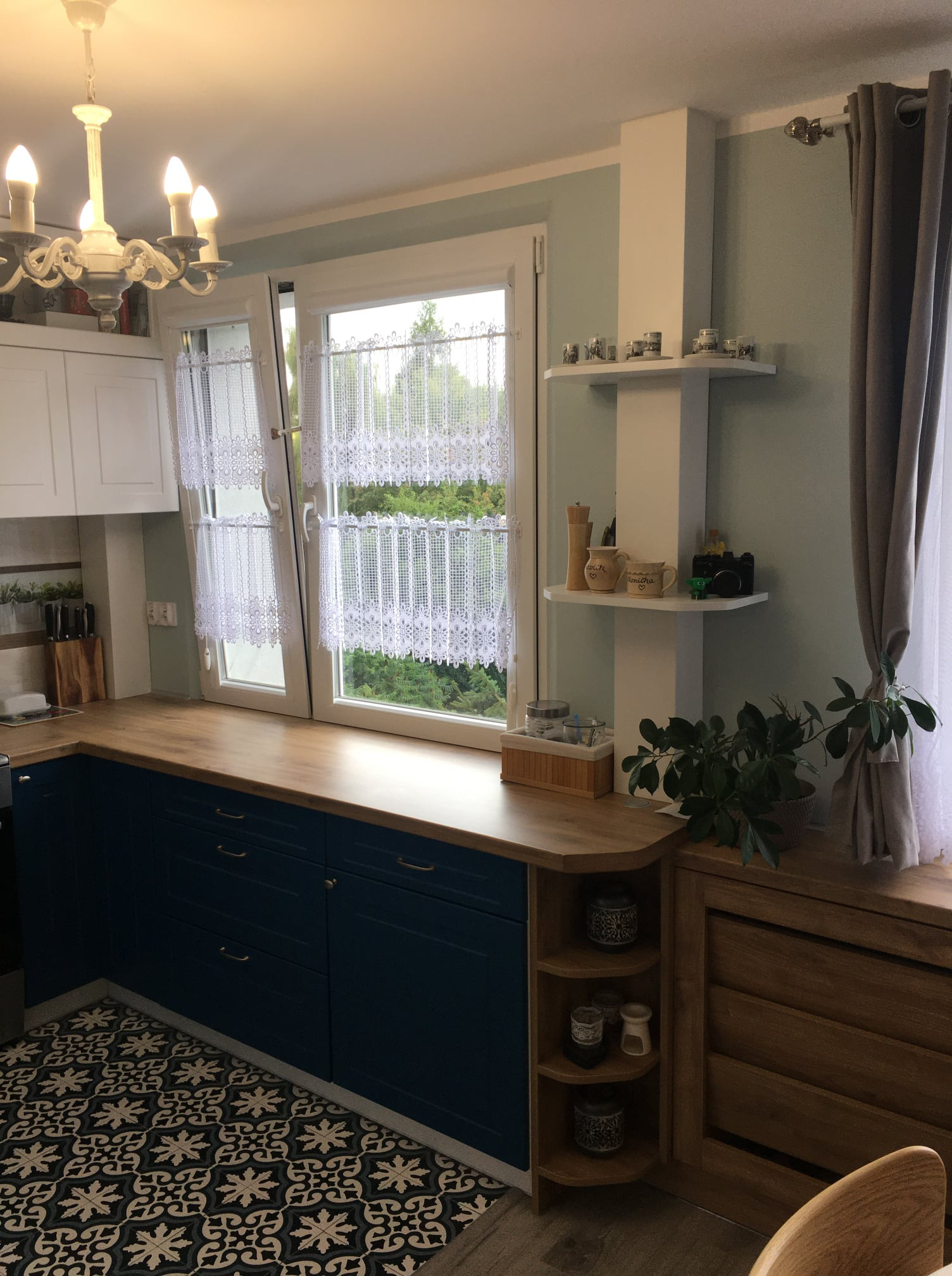 Jasna kuchnia z niebieskimi szafkami, drewnianym blatem, wzorzystą podłogą i oknem z koronkowymi firankami, uzupełniona o półki z dekoracjami i rośliną doniczkową.