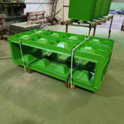 Świeżo pomalowana na zielono metalowa konstrukcja z kwadratowymi otworami, zabezpieczona białym sznurkiem, stoi na drewnianych klockach w hali produkcyjnej; w tle widać stos pomalowanych na zielono...