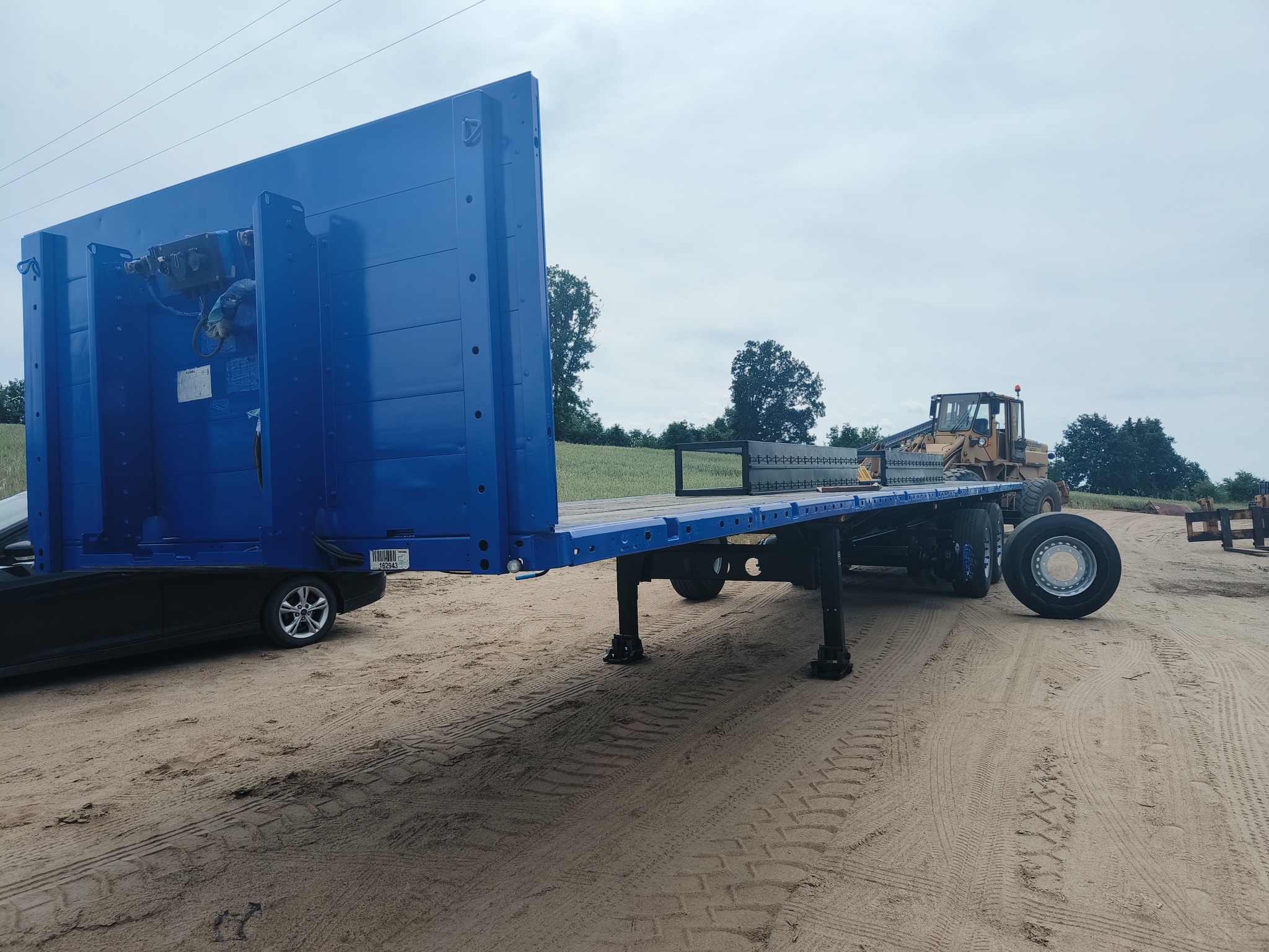 Niebieska naczepa niskopodwoziowa z widocznym ciągnikiem na piaszczystym terenie, ustawiona na podporach, z częściowo widocznym samochodem osobowym z przodu.