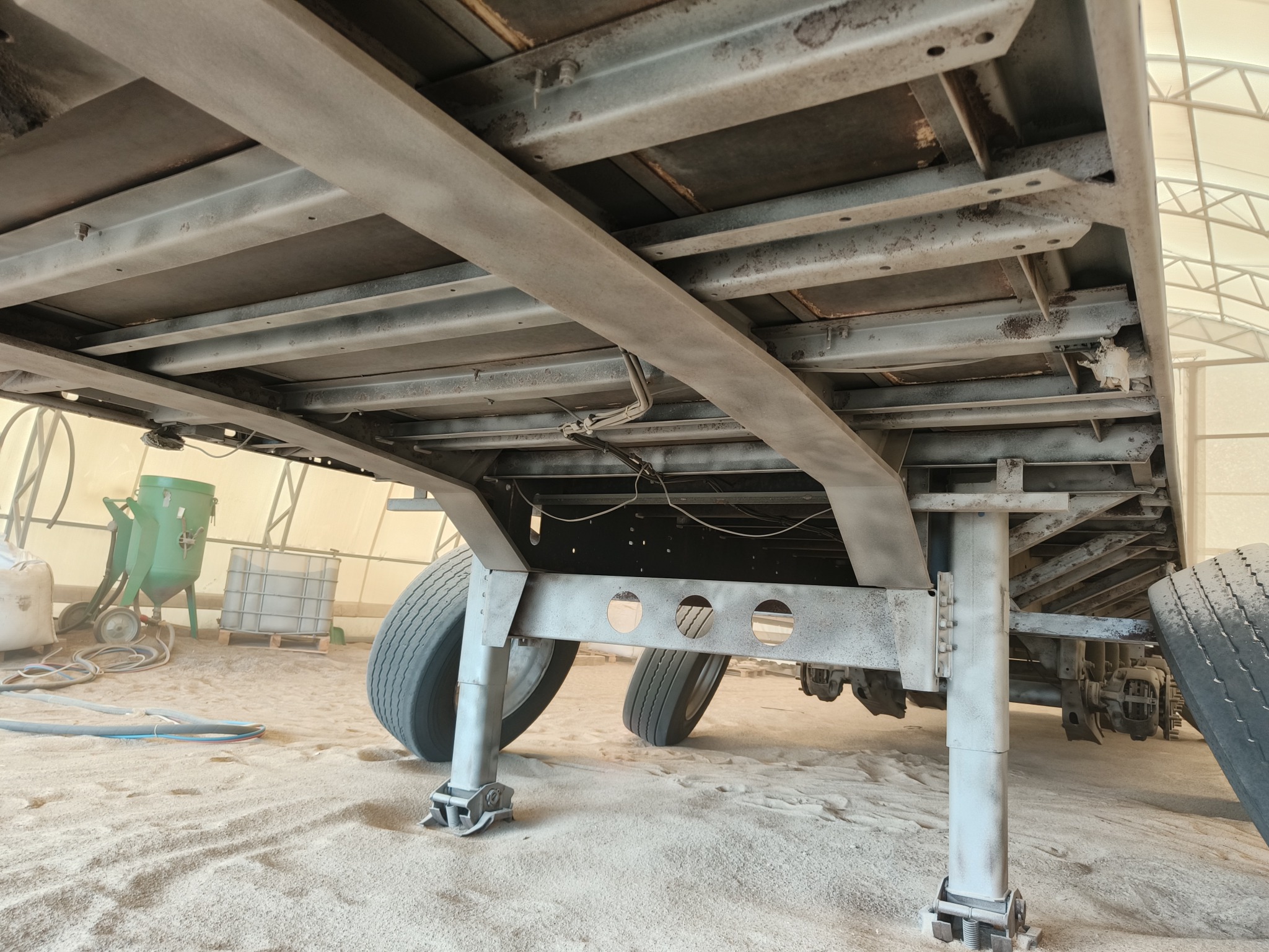 Przyczepa samochodowa po piaskowaniu, widok od spodu, ukazujący metalową konstrukcję i podwozie w piaskowanym otoczeniu warsztatowym.