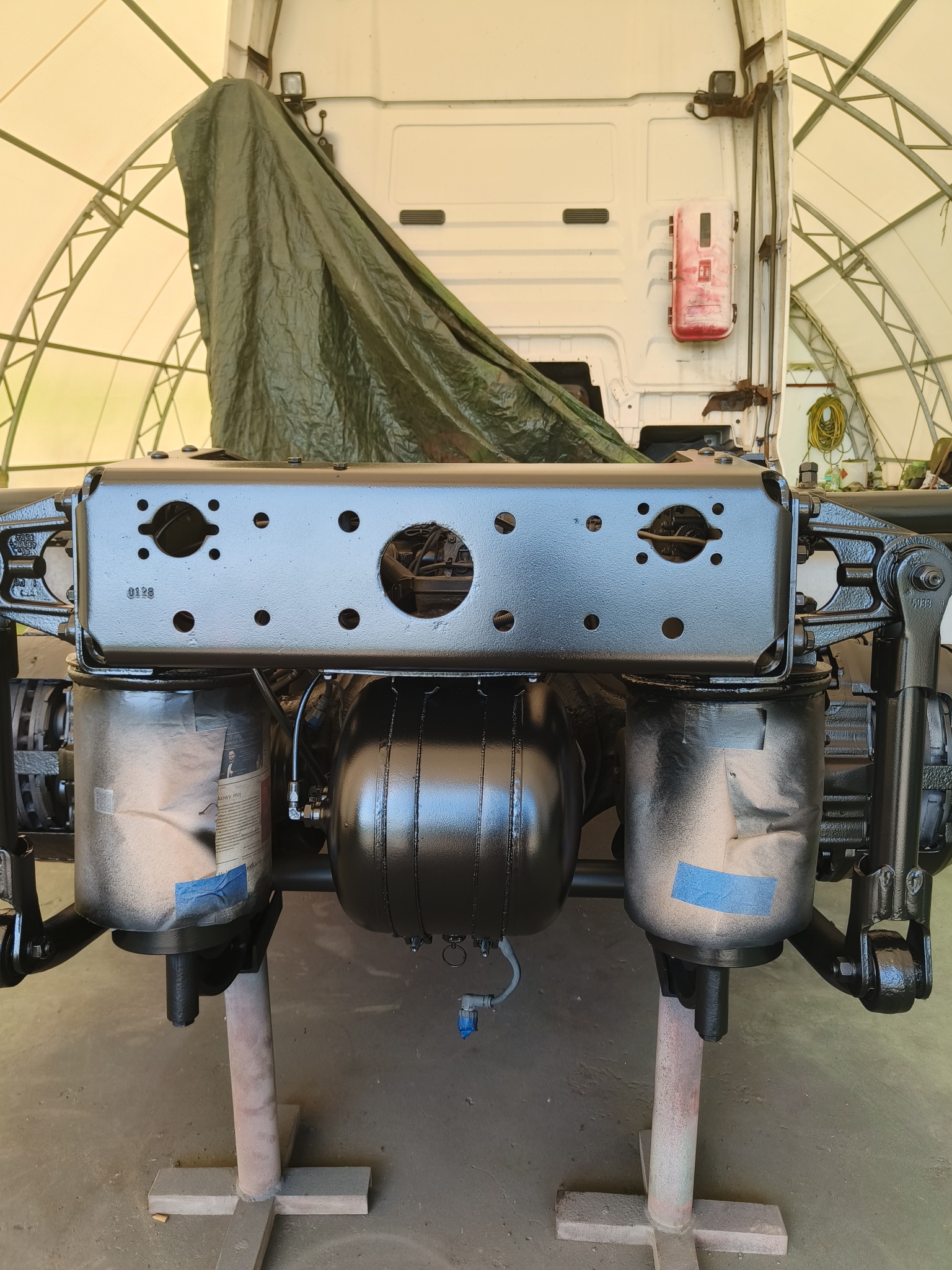 Elementy podwozia ciężarówki po renowacji, pomalowane na czarno, zabezpieczone taśmą i papierem, widoczne w warsztacie pod zadaszeniem.