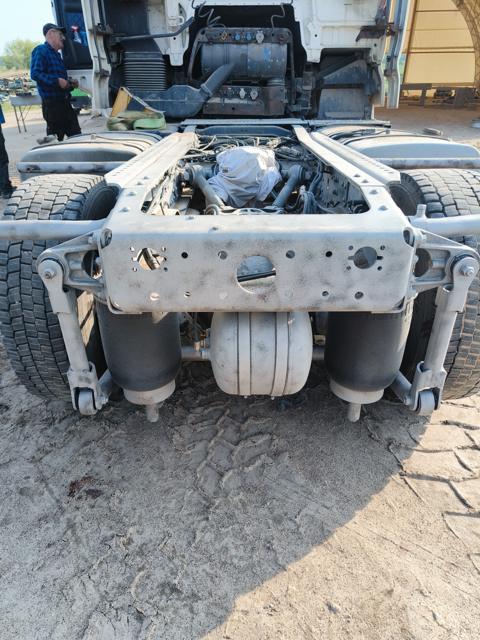 Podwozie ciężarówki z zawieszeniem pneumatycznym, widok od tyłu, z częściowo zdemontowanym silnikiem i kabiną uniesioną do góry, na piaszczystym podłożu.