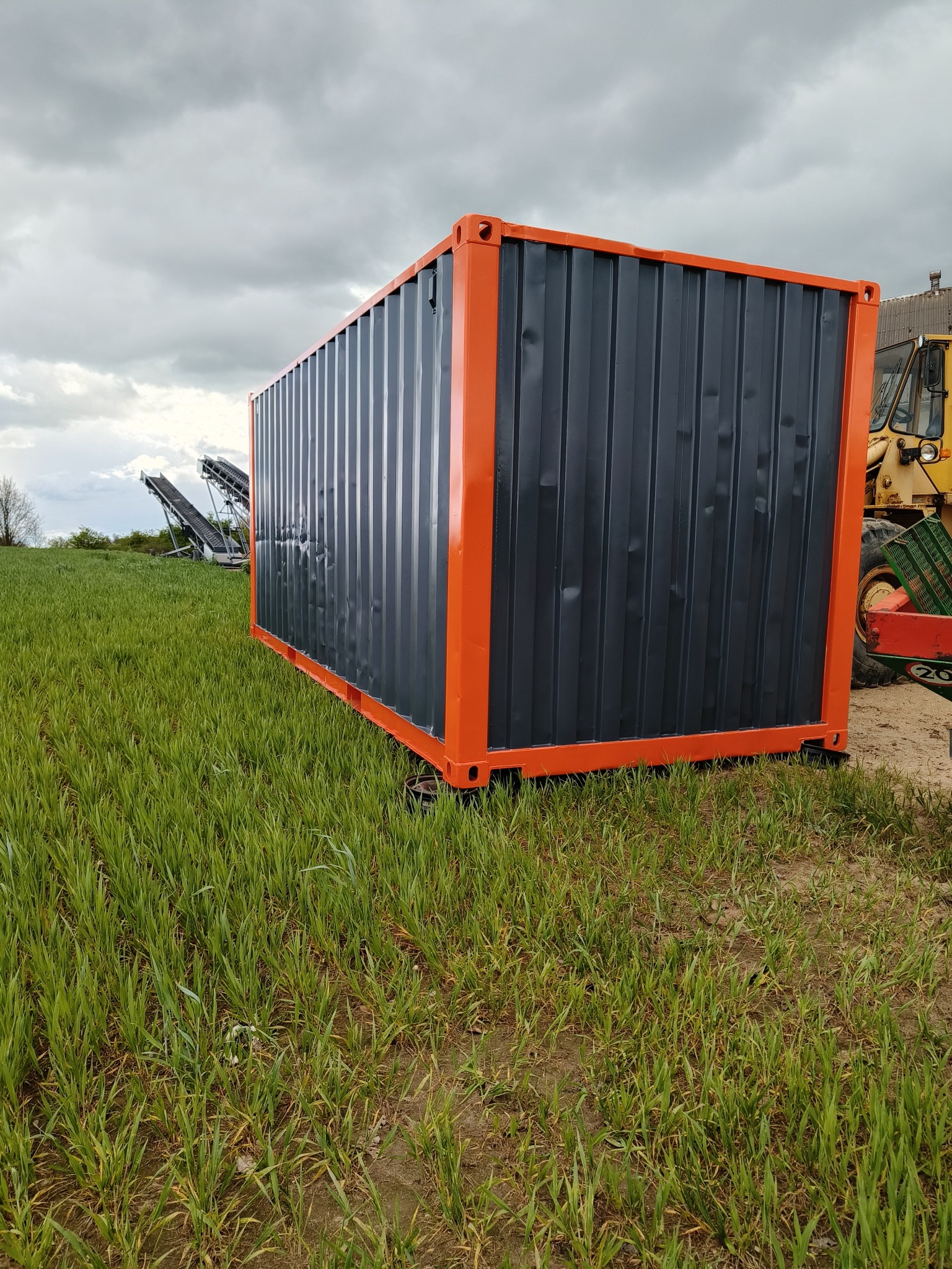 Nowy, pomalowany na szaro i obramowany na pomarańczowo kontener transportowy ustawiony na polu z młodą zieloną trawą, pod pochmurnym niebem. W tle widoczny traktor i elementy maszyny rolniczej.