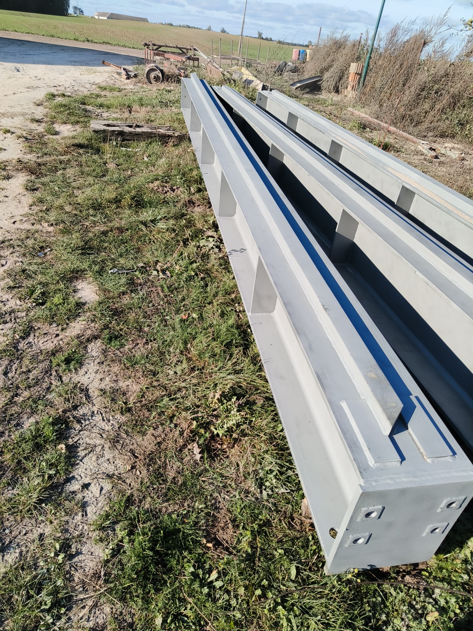 Trzy pomalowane na szaro, stalowe belki o przekroju prostokątnym, ułożone równolegle na trawie, z widocznym fragmentem przyczepy rolniczej w tle.