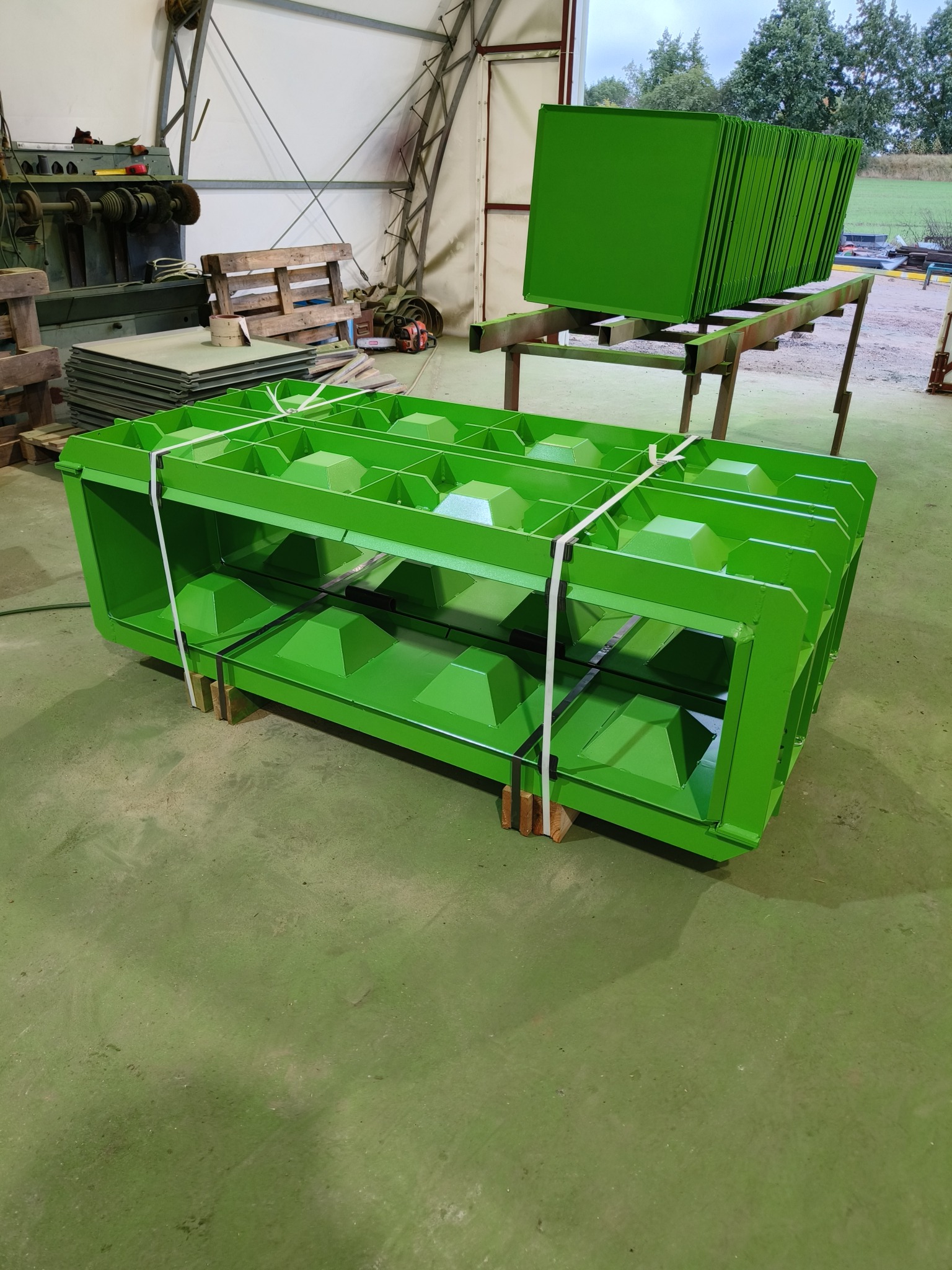 Świeżo pomalowana na zielono metalowa konstrukcja z kwadratowymi otworami, zabezpieczona białym sznurkiem, stoi na drewnianych klockach w hali produkcyjnej; w tle widać stos pomalowanych na zielono...