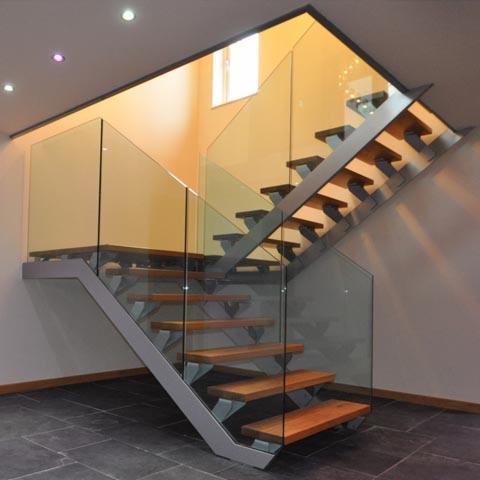 Nowoczesne schody wewnętrzne z drewnianymi stopniami, metalową konstrukcją i szklanymi balustradami, oświetlone punktowo.