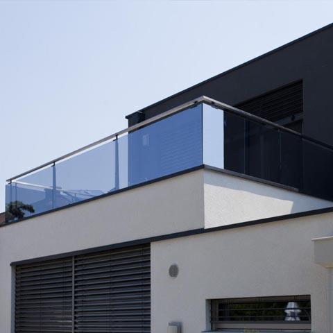 Nowoczesna szklana balustrada balkonowa z metalowymi słupkami, zamontowana na białym budynku z szarymi żaluzjami.