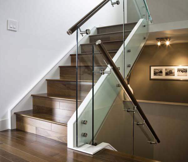 Nowoczesne schody z drewnianymi stopniami, szklaną balustradą i ciemnobrązową poręczą, oświetlone punktowo.