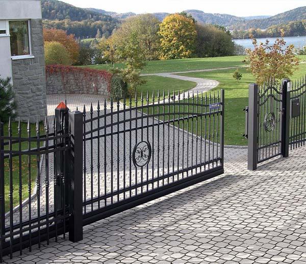 Otwarta, automatyczna brama wjazdowa z kutego żelaza, zdobiona owalnym ornamentem, na posesji z brukowaną nawierzchnią i widokiem na jezioro w tle.