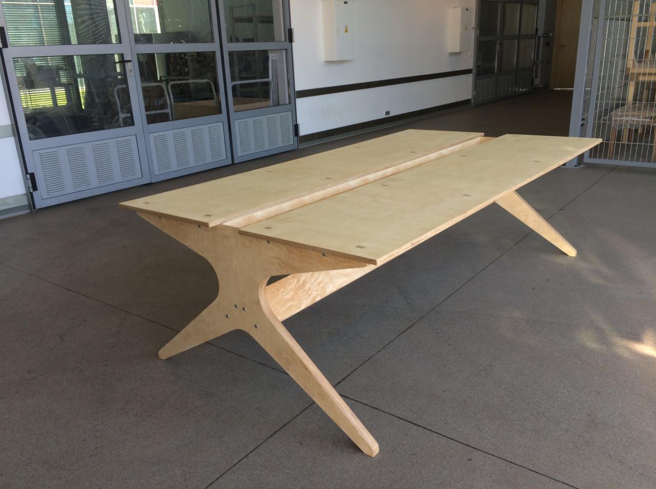 Długi, surowy stół z jasnej sklejki z nietypową podstawą w kształcie litery X, widoczne mocowania śrubami, ustawiony w przestrzeni publicznej z szarymi drzwiami w tle.