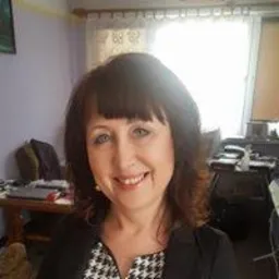 Portret uśmiechniętej kobiety z ciemnymi włosami i niebieskimi oczami, ubrana w czarną marynarkę w biało-czarny wzór, na tle biura z biurkiem i komputerem.