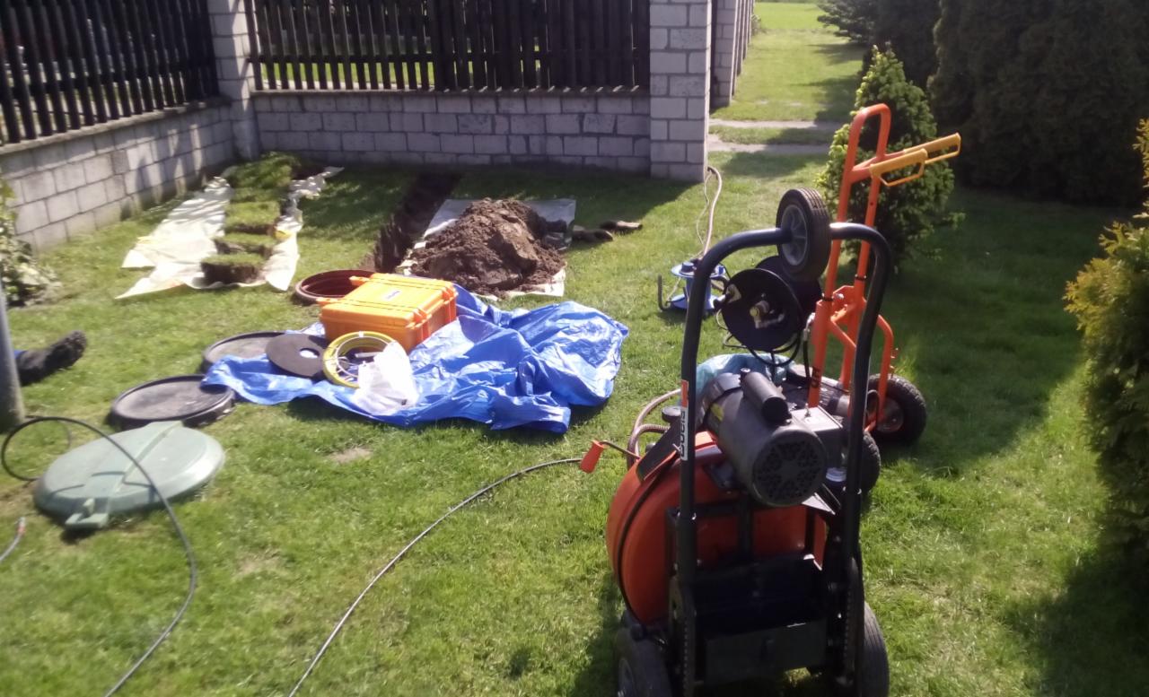 Otwarty wykop w trawie z rozłożoną niebieską plandeką, żółtą skrzynką narzędziową, pokrywami studzienek i specjalistycznym pomarańczowym sprzętem do inspekcji i czyszczenia rur.