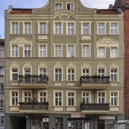 Apa Piotr Ryba Architekci - Odrestaurowana kamienica w odcieniach oliwki z kremowymi detalami architektonicznymi, balkony z ozdobnymi balustradami i widokiem na ulicę z przystankiem autobusowym.