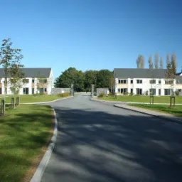 Dwa identyczne, dwukondygnacyjne domy z szarymi dachami i białymi elewacjami, symetrycznie rozmieszczone po obu stronach drogi wjazdowej, z młodymi drzewkami zasadzonymi przed nimi, widok na bramę...