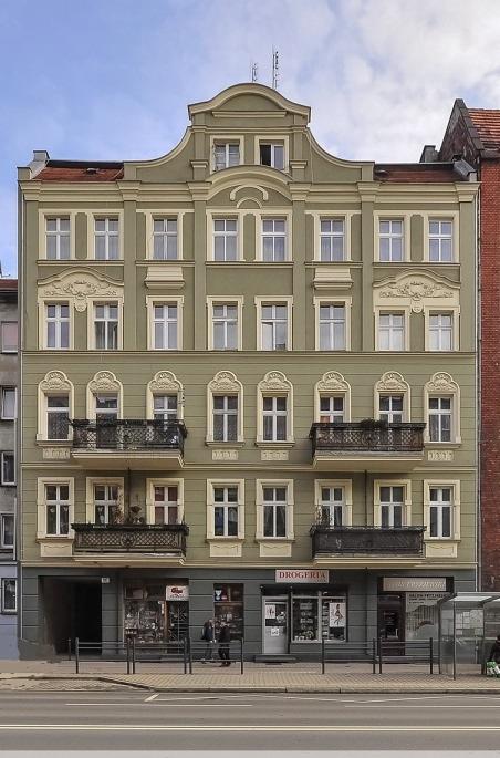Odrestaurowana kamienica w odcieniach oliwki z kremowymi detalami architektonicznymi, balkony z ozdobnymi balustradami i widokiem na ulicę z przystankiem autobusowym.