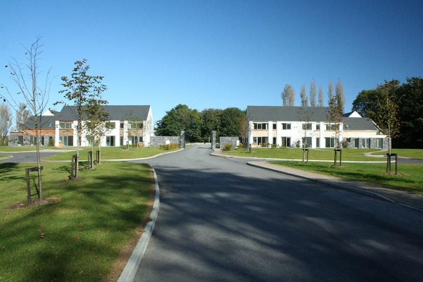 Dwa identyczne, dwukondygnacyjne domy z szarymi dachami i białymi elewacjami, symetrycznie rozmieszczone po obu stronach drogi wjazdowej, z młodymi drzewkami zasadzonymi przed nimi, widok na bramę...