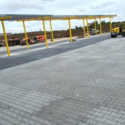 Mal-Bruk - Rozległy parking z szarej kostki betonowej ułożonej w zygzak, częściowo zacieniony konstrukcją z żółtymi słupami, z widocznym walcem drogowym i paletami z kostką w tle.