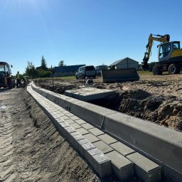 Mal-Bruk - Świeżo ułożone szare kostki betonowe tworzą krawężnik wzdłuż drogi gruntowej, z widocznymi śladami opon po ciężkim sprzęcie, w tle żółta koparka i buldożer oraz pracownicy w kamizelkach...