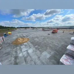 Układanie szarej kostki betonowej na dużej powierzchni w Domanicach, widoczne palety z kostką, piasek, ekipa budowlana i samochód dostawczy.