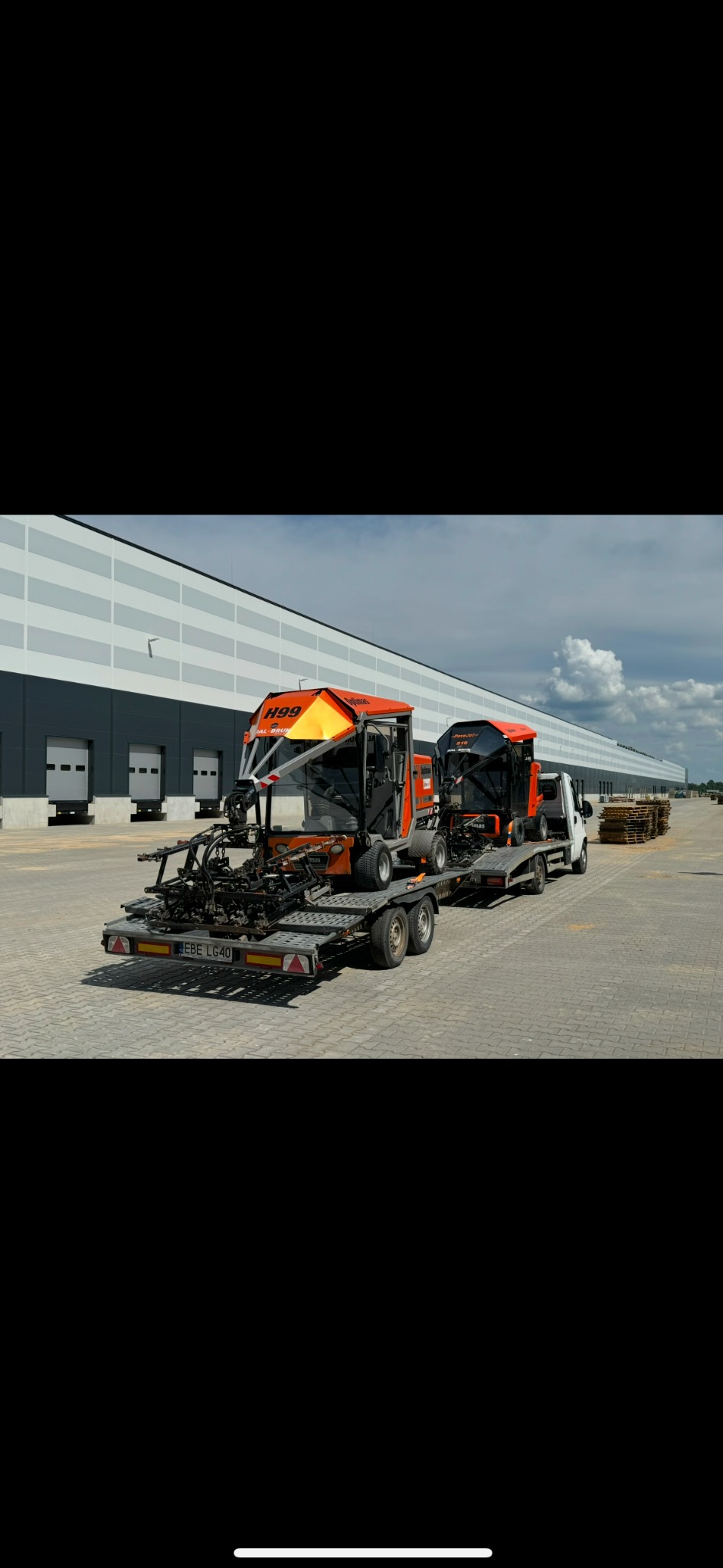 Dwa pomarańczowe, specjalistyczne pojazdy do układania nawierzchni przewożone na przyczepie lawetą, na tle magazynu z rampami załadunkowymi.