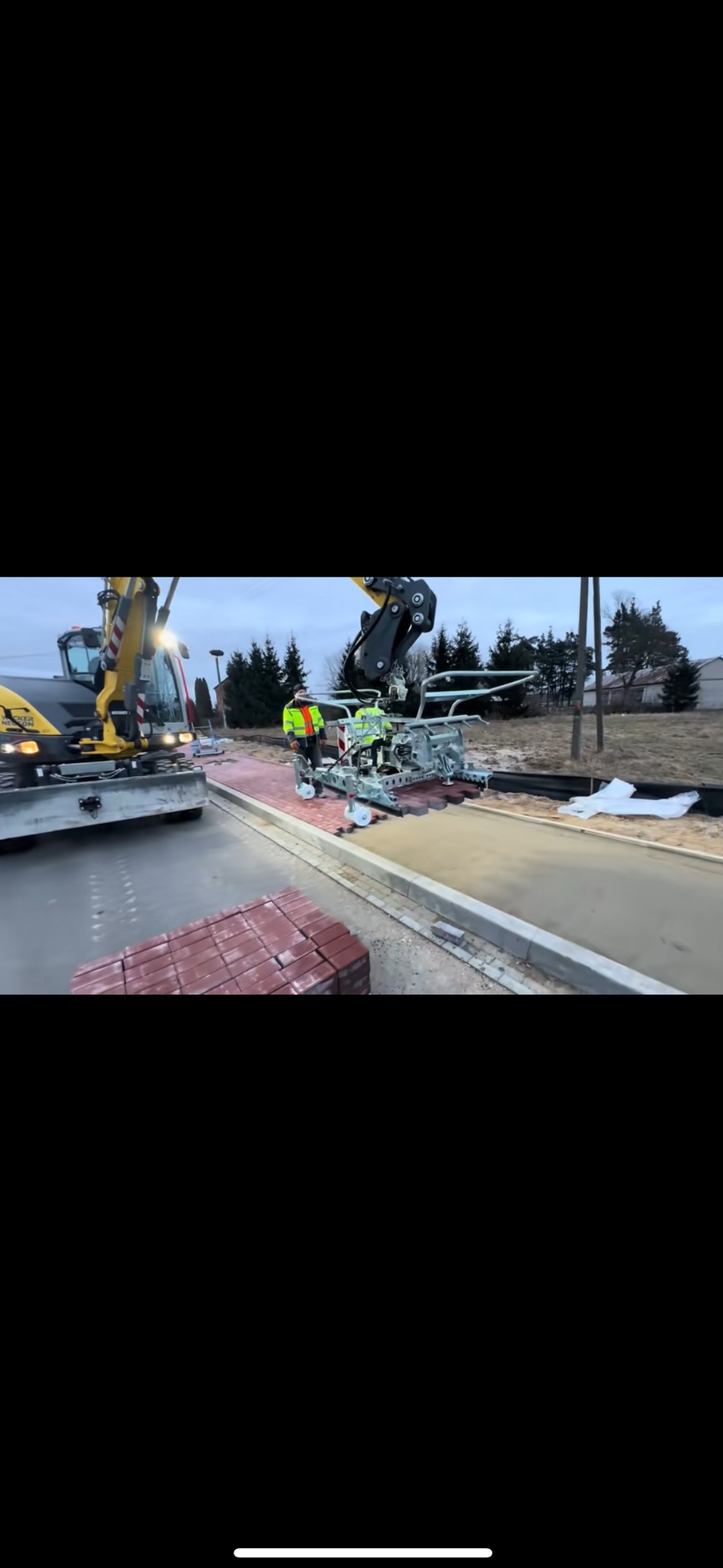 Automatyczne układanie czerwonej kostki brukowej za pomocą specjalistycznej maszyny, z udziałem operatora w odblaskowej kamizelce, w tle widoczny mini-koparka i teren budowy.