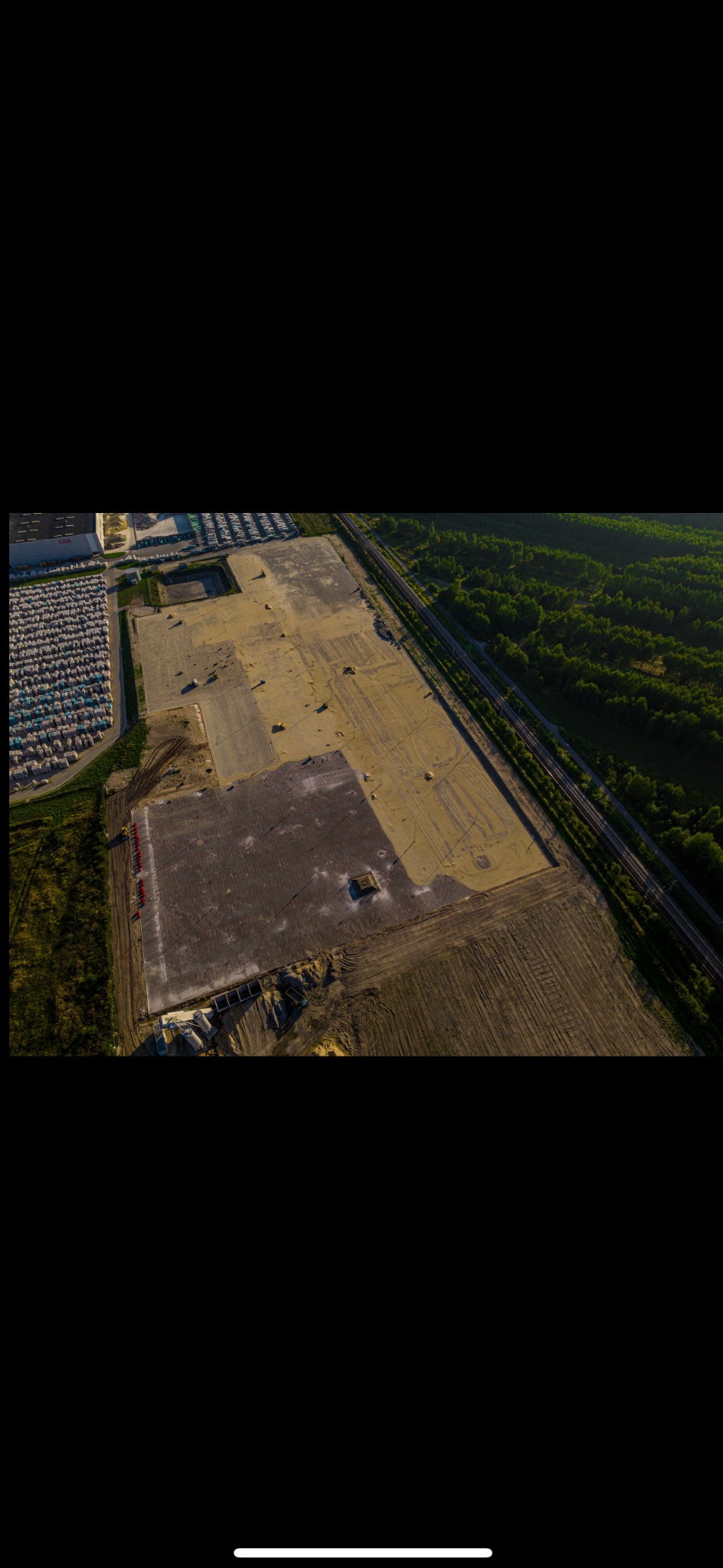 Zdjęcie z drona przedstawia teren budowy z przygotowanym podłożem, składowiskiem kostki betonowej i fragmentem lasu w tle.