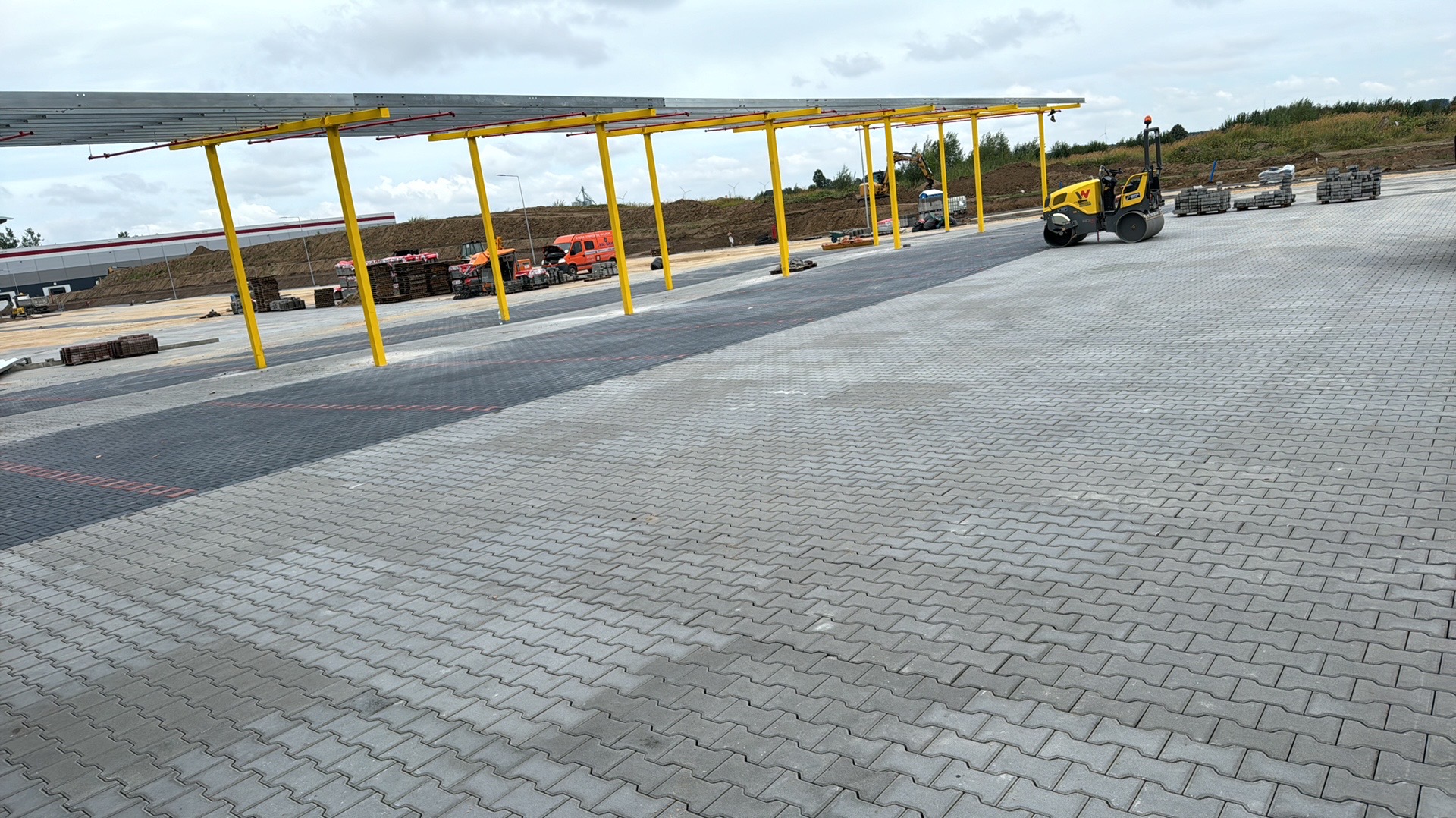 Rozległy parking z szarej kostki betonowej ułożonej w zygzak, częściowo zacieniony konstrukcją z żółtymi słupami, z widocznym walcem drogowym i paletami z kostką w tle.