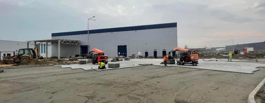 Układanie szarej kostki brukowej na terenie przemysłowym, widoczne maszyny do układania, koparka i pracownicy w odblaskowych kamizelkach na tle hali magazynowej.