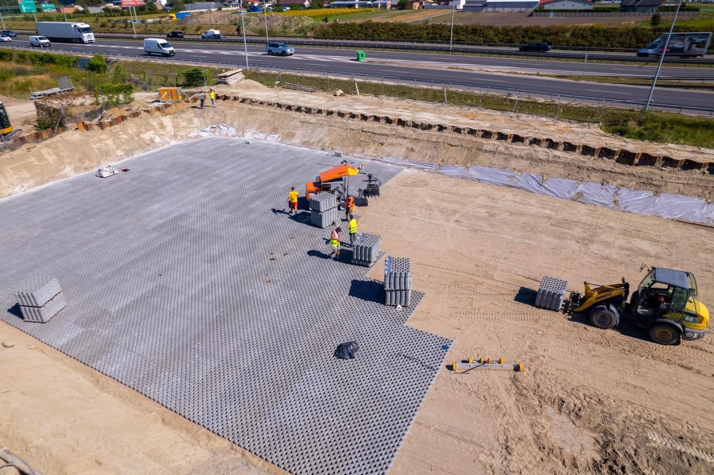 Z góry widok na plac budowy z ułożoną częścią ażurowych płyt betonowych, pracownicy w żółtych kamizelkach, mała ładowarka z kostką, w tle ruchliwa droga ekspresowa.